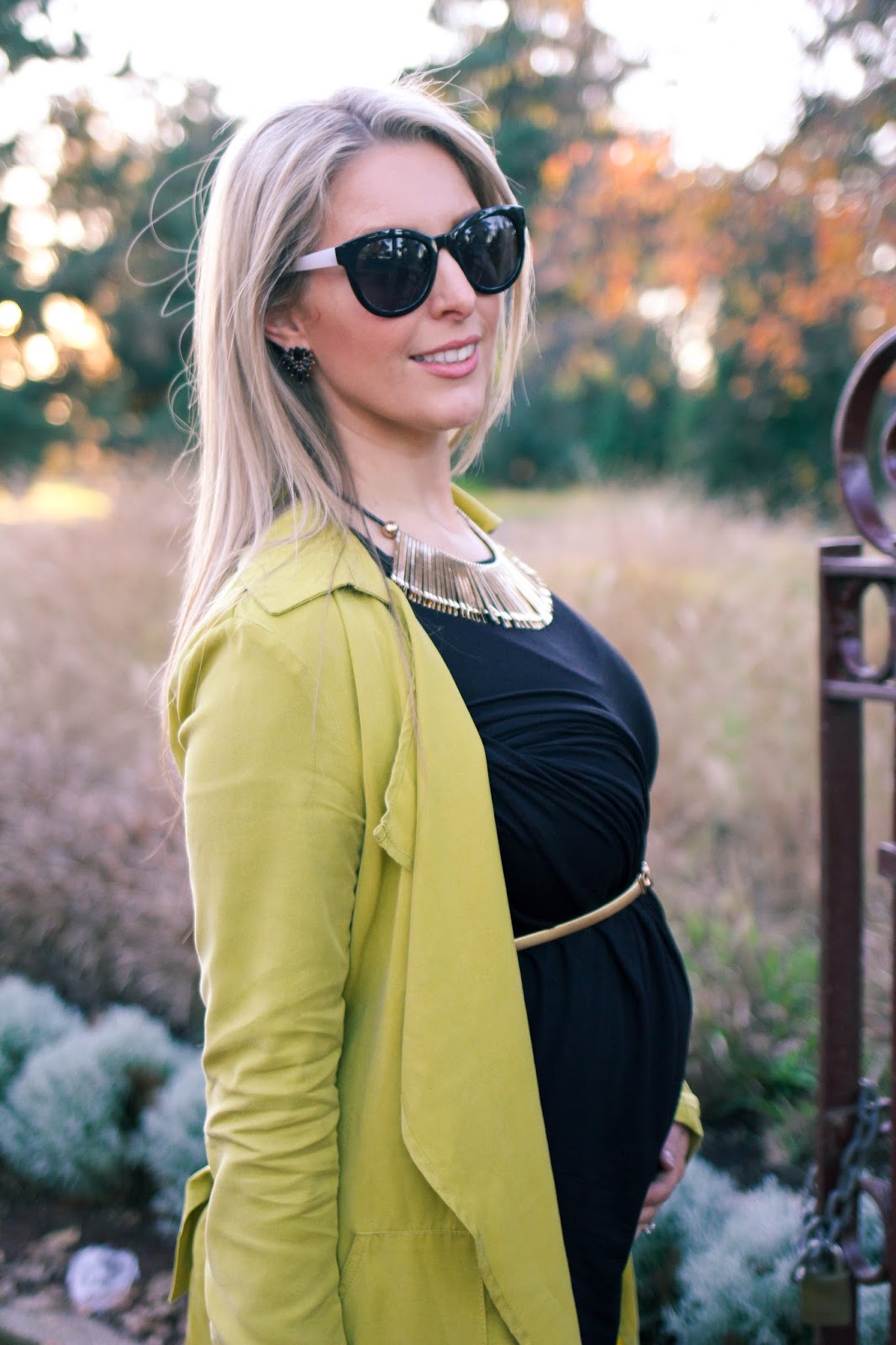 The Goldfields Girl styling maternity fashion. She wears black maternity dress, green jacket, statement gold necklace, brown boots and Michael Kors bag.