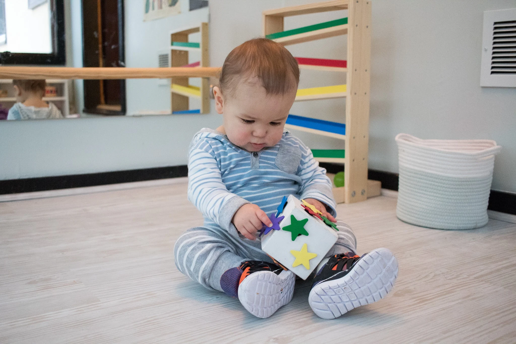 An older baby sits in his Montessori playroom using a DIY velcro block covered in wooden stars. He looks down using fine motor skills to pull one of the stars from the block as he plays with the Montessori friendly DIY.