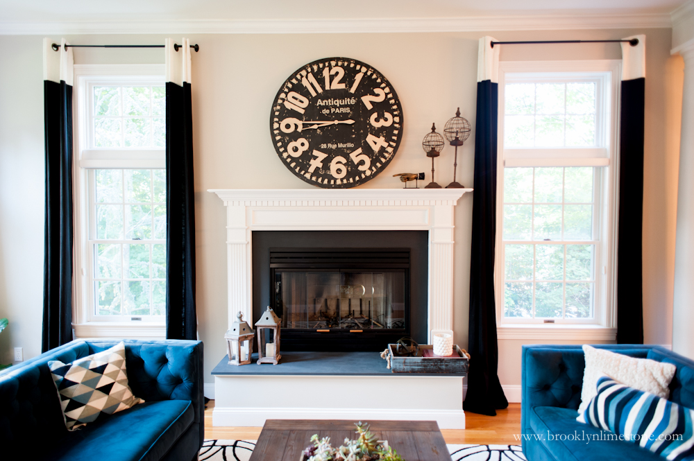 Exterior wall with fireplace that now has windows with black and white solid curtain panels on them, 2 teal sofas that face each other flank either side of the fireplace