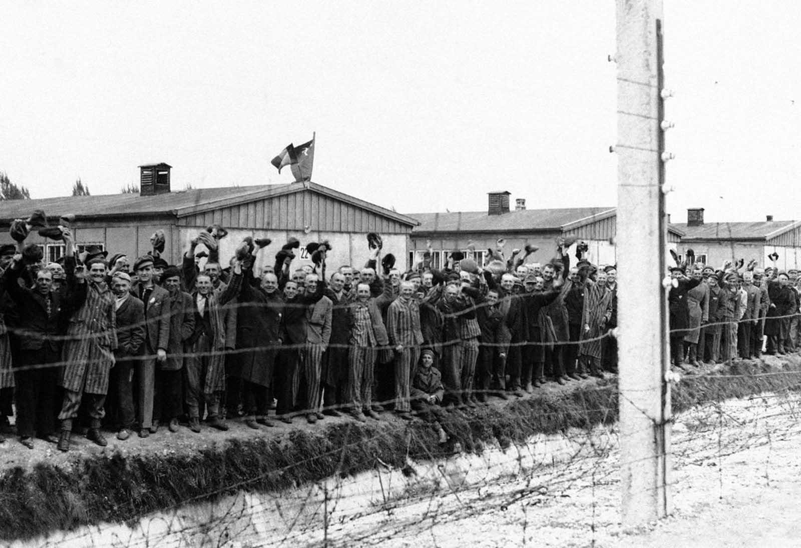 A Dachau koncentrációs tábor elektromos kerítésén tartózkodók ragaszkodnak a német Dachauban, az amerikai katonákhoz egy korábbi fotón.  Néhányuk csíkos kék és fehér börtönruhát visel.  Kunyhóikat minden nemzet zászlóival díszítették, amelyeket titokban készítettek, amikor hallották, hogy a 42. szivárványosztály fegyverei egyre hangosabbak lettek a Dachau-hoz való megközelítésben.