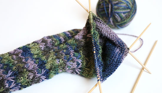 Hand knit wool socks in progress on bamboo needles in blues, purples and greens on a white background.