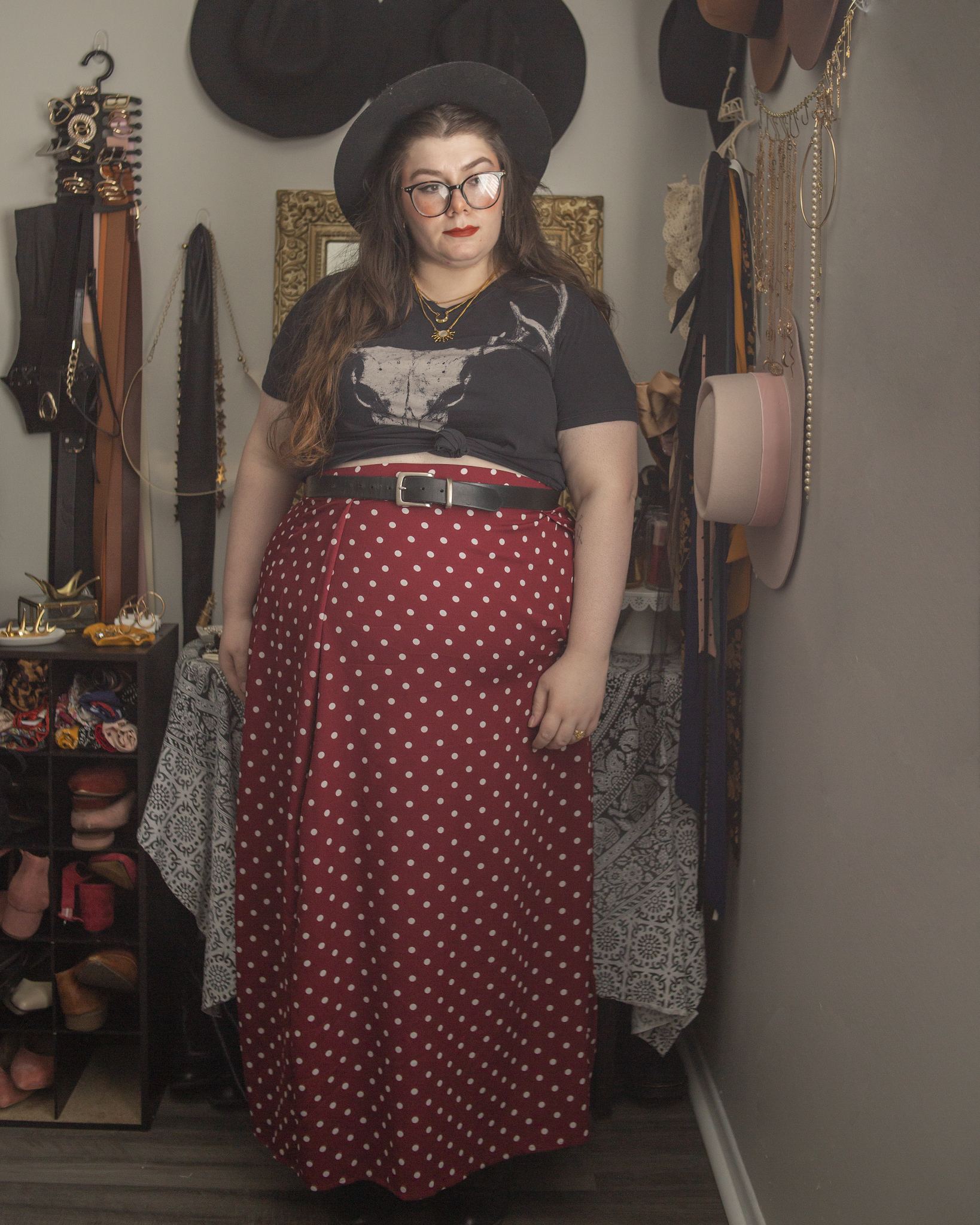 An outfit consisting of a black v-neck t-shirt with a bone colored deer skull motif, tied in a knot, with a white on maroon red large polka dot midi skirt with a high slit, and heels.