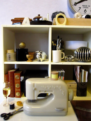 1/12 scale sewing machine on a work table surrounded by scissors, thread a tape measure, cutting board, ruler and glass of wine. Behind it is cube shelving full of books, ornaments and stationery supplies.