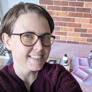 Sarah, 30 year old white woman, taking a selfie smiling awkwardly at the camera, behind her is a dining table with disassembled photo frames and paints.