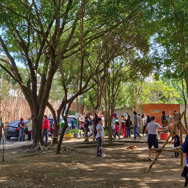 pessoas interagindo no espaço da praça, um local com árvores e brinquedos de bambu.