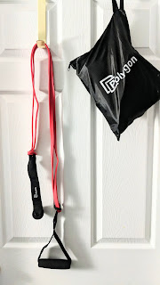 A Polygon resistance band with attached hand and door anchor hanging on a hook on a door. To the right is the Polygon case filled with the remaining resistance bands, also hanging on the door.