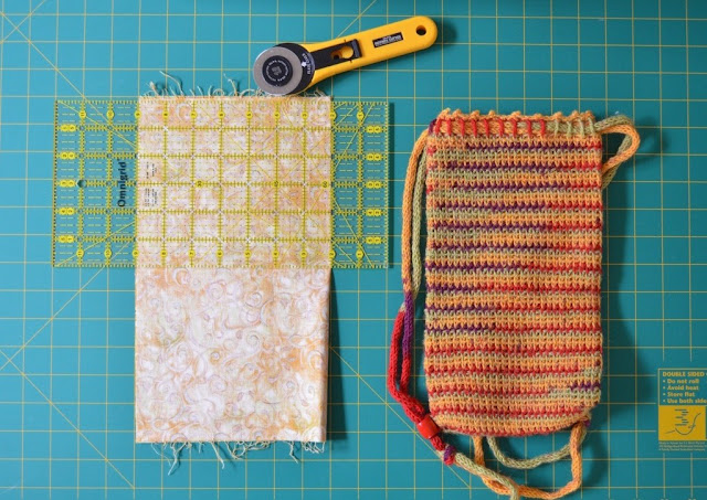 The folded fabric is laid out on the grid next to the bag to compare heights.  The fabric is lined up with the grid and the quilters rule is aligned across the top of the fabric and with the grid. The rotary cutter is ready to trim the top edge of the fabric.