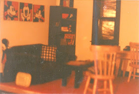 1/12 scale modern miniature scene of a lounge in a 1980s-style apartment. The carpet is red,  the walls white and the windowframes, blinds, sofa, coffee table and shelving unit black. There is a Mickey Mouse triptych above the sofa, a walkman on the coffee table and a turntable and records in the shelving unit.