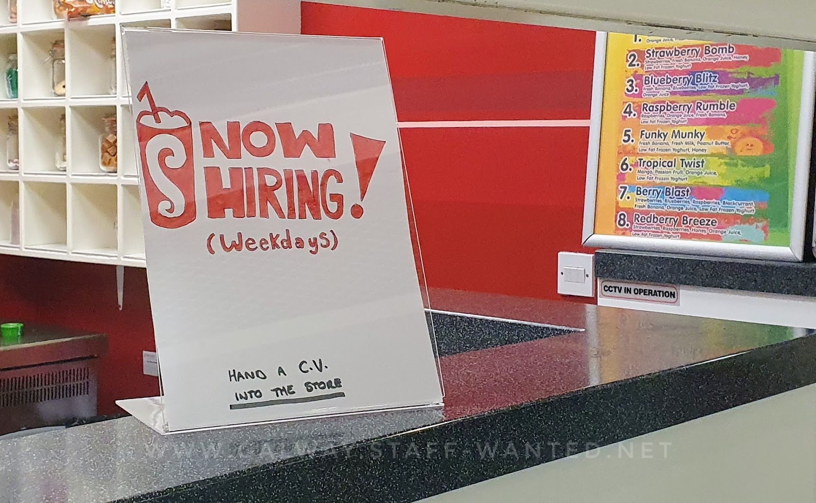 Shop counter with staff-wanted advert in a plastic display:  advertisement features a hand-drawn milkshake with a curly S and a straw.    Background includes a Shake Milkshakes menu, with drinks including Strawberry Bomb, Blueberry Blitz, Raspberry Rumble, Funky Munkey, Tropical Twist, Berry Blast and Redberry Breeze.  Sign shows ingredient list of each of these.