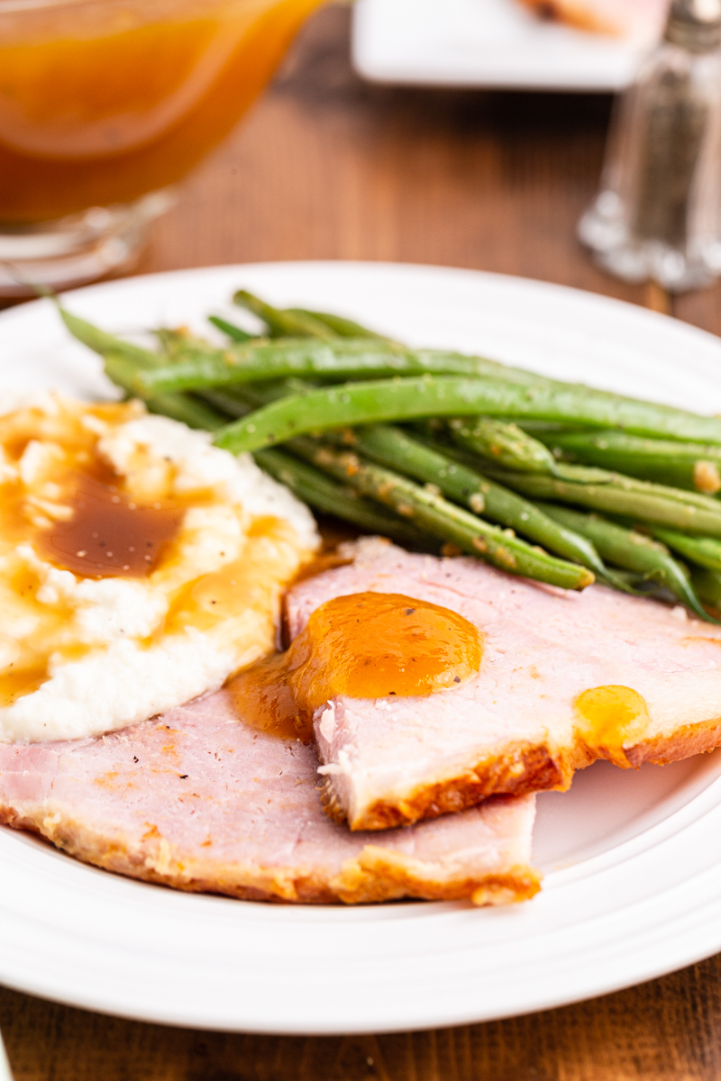 Close up photo of slices of Keto Apricot Bourbon Glazed Ham on a white plate, topped with gravy and mashed cauliflower and green beans on the side.