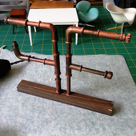 One-tweltfh scale modern miniature pipe shelving mounted on a length of wood , on a workbench next to a drill.