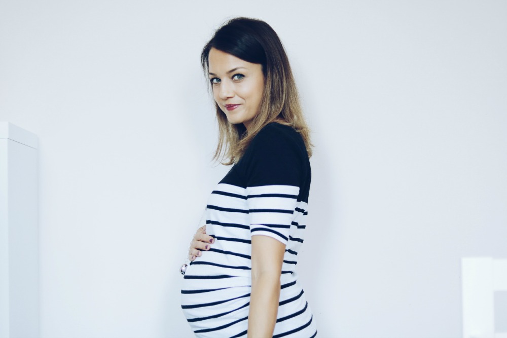 20 weeks pregnant woman in 30s with dark brown hair wearing black and white stripey top holding bump with one hand and smiling