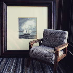 Small framed watercolour of a boat propped up against the wall. In front of it is a piece of carpet tile and a 1/12 scale modern miniature armchair in greys and browns.