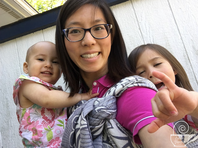 [Image of me, a light tan skin bespectacled Asian woman in a pink dress, with a preschooler making a silly face on my back in a double hammock with a candycane chess belt in a grayish purplish geofloral woven wrap. I'm holding a wiggly toddler in front who's wearing a mama-made pink floral sundress. We're outside in the shade of a 90F sunny day.]