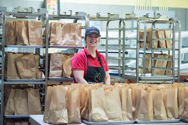 Image of a school food service employee surrounded by brown paper bag lunches