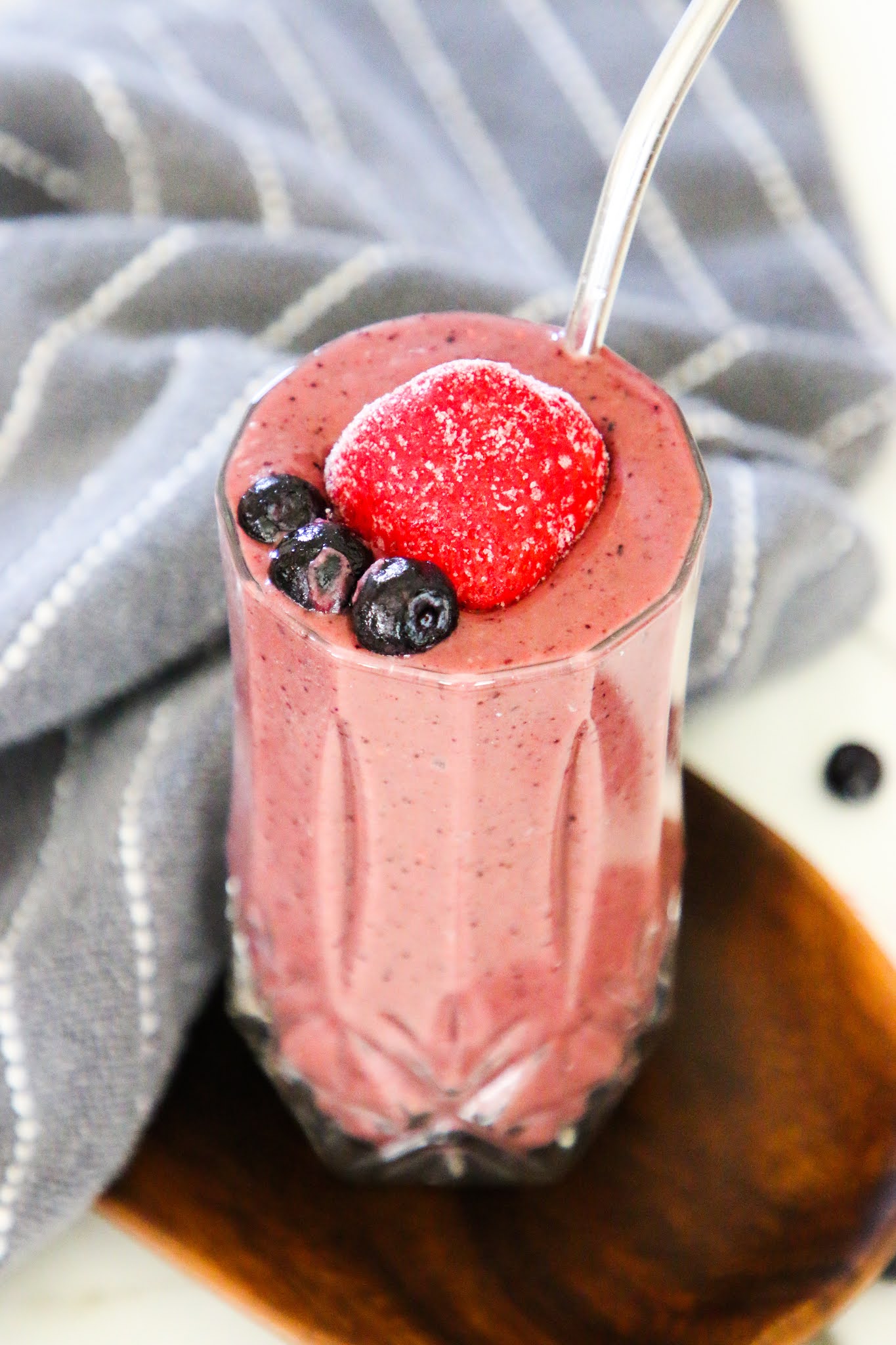 Smoothie with a big strawberry on top, sitting on a wooden platter, with a dish towel in the background.