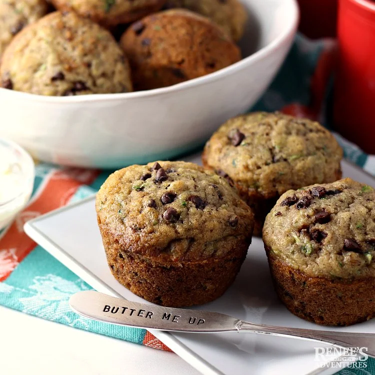 Chocolate Chip Zucchini Muffins by Renee's Kitchen Adventures in a bowl in the background with three muffins in the foreground on a white plate with a spreader for butter resting on the plate