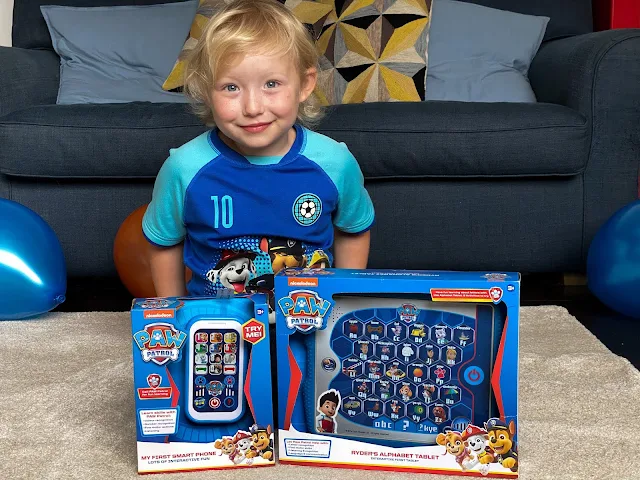 A child in PAW Patrol top sitting behind the PAW Patrol Smart Phone and Alphabet Tablet in their packaging before reviewing them