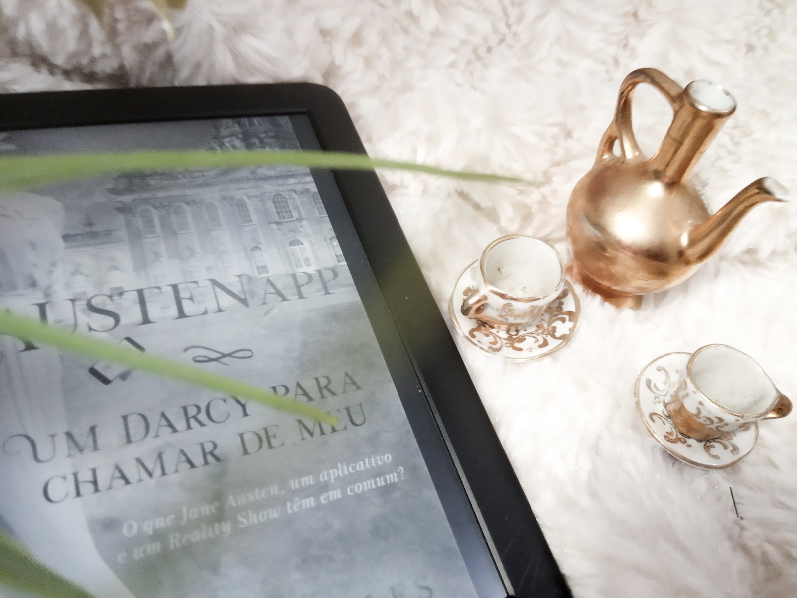 AustenApp Um Darcy para chamar de meu de Katherine Salles Foto com aparelho kindle focado na capa de AustenApp em que aparece os dizeres Um darcy para chamar de meu, ao lado direito ha miniaturas de xicaras, pires e um bule de chá sob um tecido branco e peludo