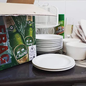Benchtop with a red, gold and green beer carton on top of it, opened, with a length of 'fragile' tape on the side. Next to the carton are two stacks of white plates, two ramekins, a glass of sparking wine, a pile of used packing paper and a can of oven cleaner.