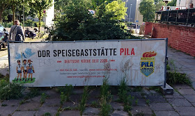 Plakat der "DDR-Speisegaststätte Pila" mit verfremdeten FDJ-Symbol und drei gezeichneten Figuren in einer Art Pionieruniform. 
