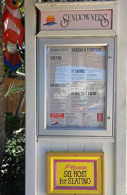 Sundowners Menu The menus is displayed outside in a glass case. A sign hangs below that reads, "Please See Host for Seating." A clown fish flag hangs from a tree in background.