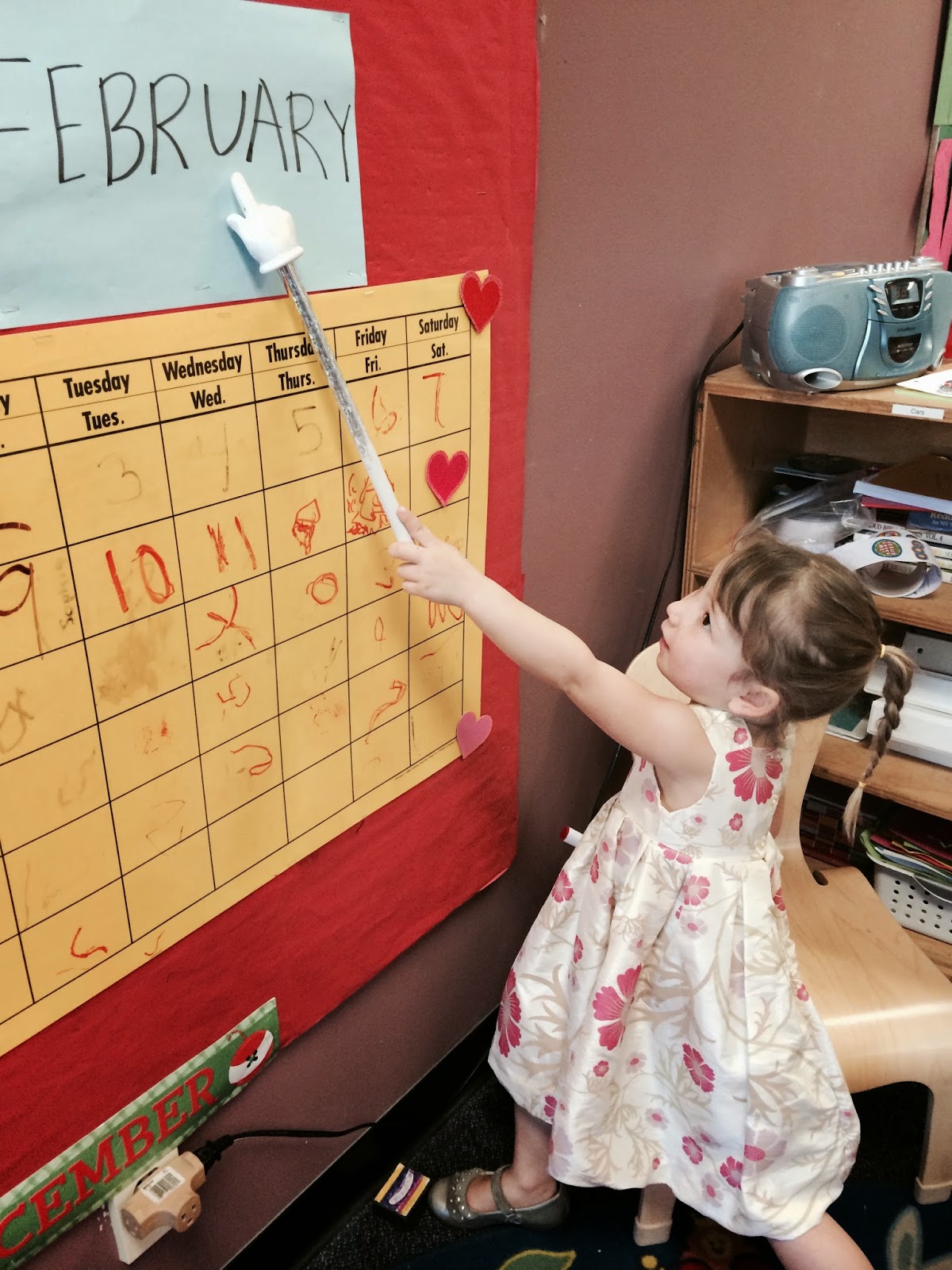 Miss E, in a floral dress, using a hand pointer (with a pointing, white hand on the end) to point to the word "February" on the preschool calendar