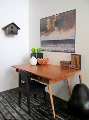 Modern miniaturecorner of a room with a mid-century modern wooden desk and chair, a large pear on the floor and a birdhouse on the wall.