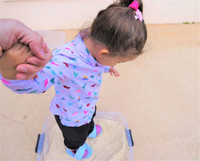 Mom hold daughter's hand as she makes deer footprints in a sandbox with her foam shoes.