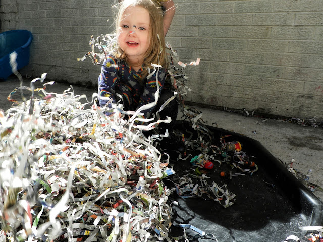 A small girl is sat behind a pile of shredded paper strips while more float around her in the air. She is blonde haired with blue eyes smiling into the canera. She is wearing a long sleeved navy dress with rainbows and elephants printed all over it. There is a white brick wall in the background.