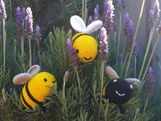 Three crocheted bees' faces poking out from between the sprigs of a lavender bush. A triangular arrangement, the top bee has a yellow face with white wings, the bottom left is sideways with a yellow face,two black stripes and silver wings, facing the top bee. Bottom right is a black-faced bee with white mouth and eyes and silver wings. Each bee is smiling.