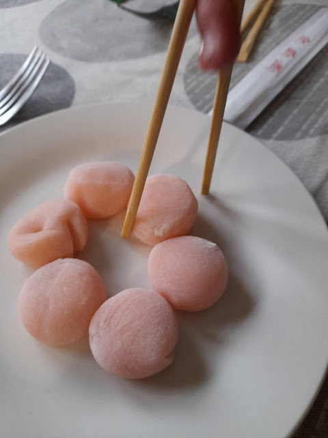 6 pink Mochi in a circle on a small white plate with a pair of chop sticks about to lift one off the plate on the right