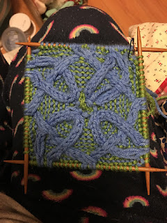An in progress blanket square on a set of double pointed needles. It has a blue cable knit knotwork on a mixed blue and green background.