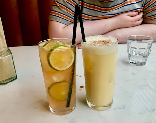 A tall glass filled with light orange liquid with two green circular discs of lime with a cylindrical black straw next to a glass filled with light yellow liquid with a black cylindrical straw on a bright background