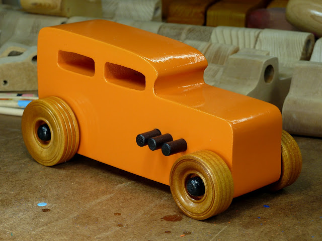 Handmade Wooden Toy Car Hot Rod 1932 Ford Sedan From the Hot Rod Freaky Ford Series Orange & Black Wooden Toys In the Background On Workbench