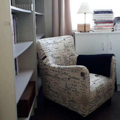 Reading chair in fawn, with black writing, in the corner of a library, next to a white cupboard under a window.
