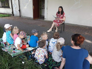 Pani bibliotekarka siedzi na krześle i czyta przedszkolakom bajkę. Dzieci z panią przedszkolanką siedzą na kocu na trawie.