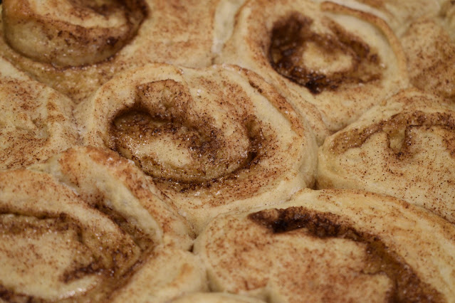 A photo of about 7 light-beige cinnamon rolls sitting in a pan (the pan is not visible in the frame). The cinnamon rolls are covered in reddish-brown specks of cinnamon and have a slightly glossy appearance.