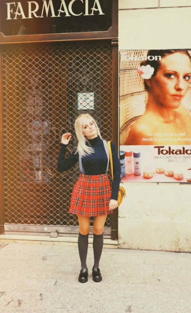 Alejandra Colomera vestida con jersey azul, falda roja cuadros escoceses, tirantes en color gris, calcetas gris y zapatos estilo colegial negro frente a una farmacia antigua