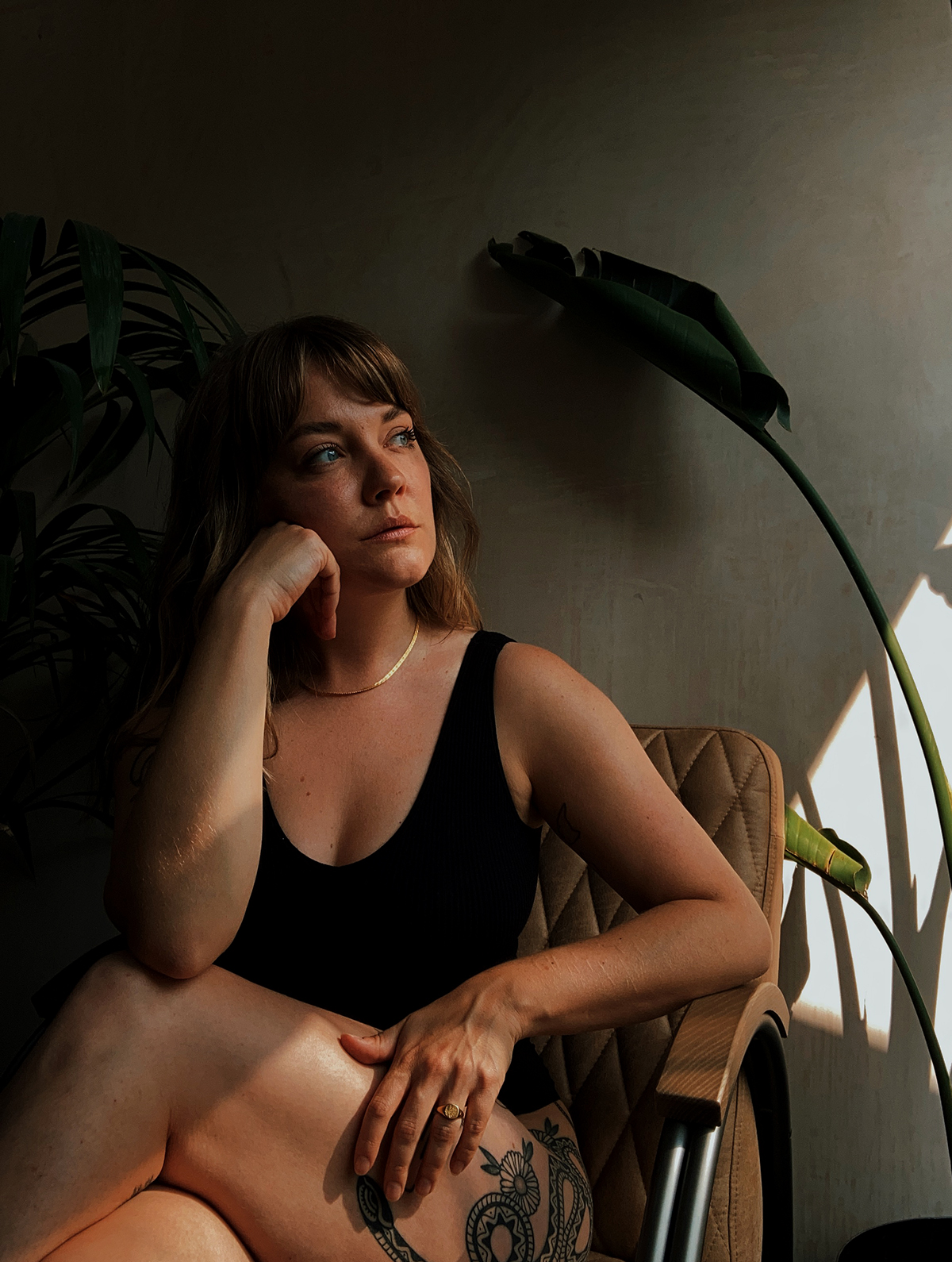 Lyzi sitting on a vintage chair in front of a plaster wall and large plants. Wearing simple black underwear and a Daisy x Estee Lalonde snake chain and forget me not signet ring.