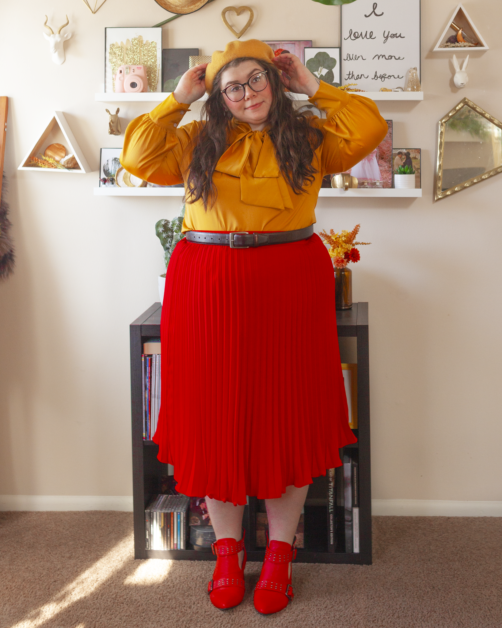 An outfit consisting of a mustard yellow beret, a silk mustard yellow blouse with a connected pussy bow tied into a large bow around the neck, tucked into a red pleated midi skirt and red boots.