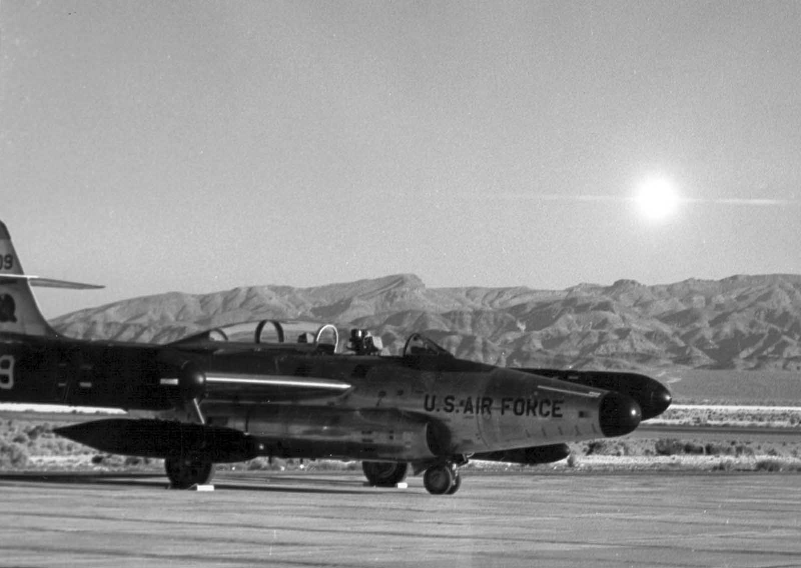 The flash of the exploding nuclear warhead of an air-to-air rocket is shown as a bright sun in the eastern sky at 7:30 a.m. on July 19, 1957 at Indian Springs Air Force Base, some 30 miles away from the point of detonation. A Scorpion, sister ship of the launching aircraft, is in the foreground.