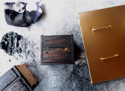 Miniature drawers being stained a dark colour, with a puddle of stain and a staining cloth next to them. On the other side are 4 drawer handles for the drawers, spraypainted gold.