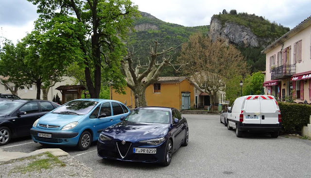 Blaue Alfa Giulia im Vordergrund neben anderen Autos geparkt mit Kalkbergen im Hintergrund und Häuser rechts davon