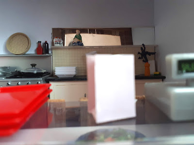 1/12 scale modern miniature takeaway food counter with trays, till, and glass full of cutlery on top of the counter. Behind, in the kitchen is a stove with pots, sauces above and a bench with plates, and ornaments above.