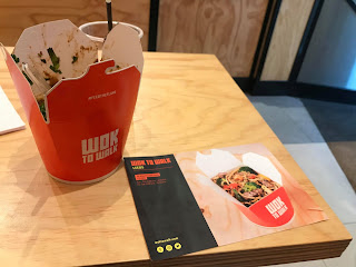 A red square cardboard take out box with wok to walk in white font filled with bright golden noodles,,vegetables and meat next to a black rectangular piece of paper on a rectangular white table on a bright background 