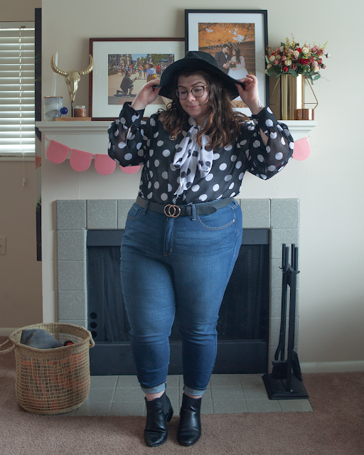 An outfit consisting of a wide brim black hat, a black and white blouse dotted blouse with a contrasting white on black dotted neck tie tied in a bow, tucked into high waist blue denim jeans and black chelsea boots.