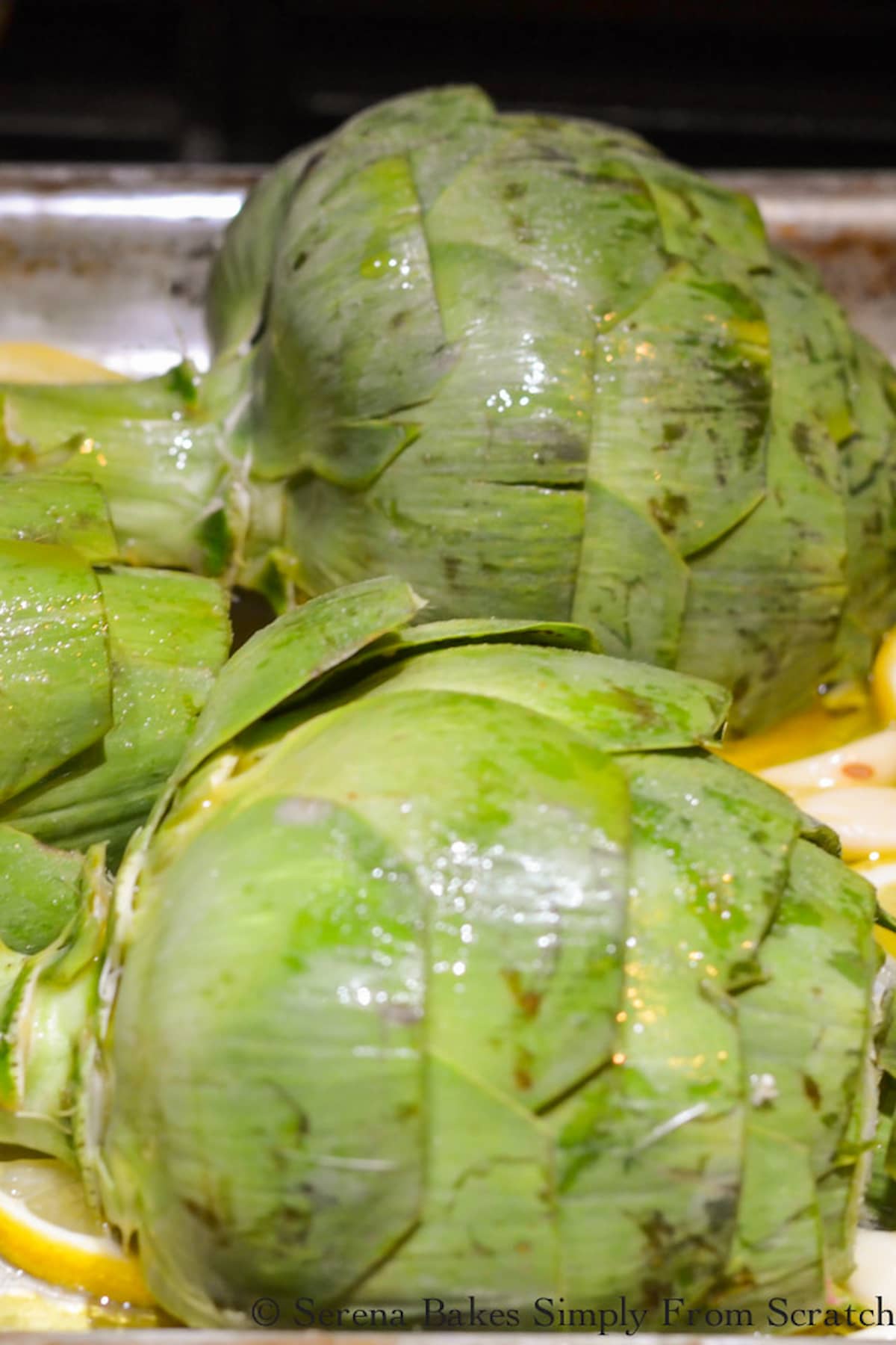 Uncooked Artichokes placed cut side down on a baking sheet drizzled with olive oil and lemon slices.