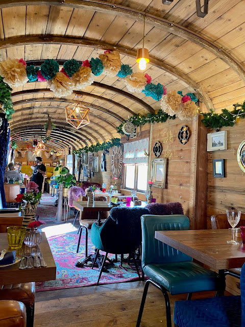 The interior of a railway carriage. The walls are wooden, and decorated with pictures and mirrors. The carriage is filled with chairs and dining tables, with an eclectic range of vases and glassware on them