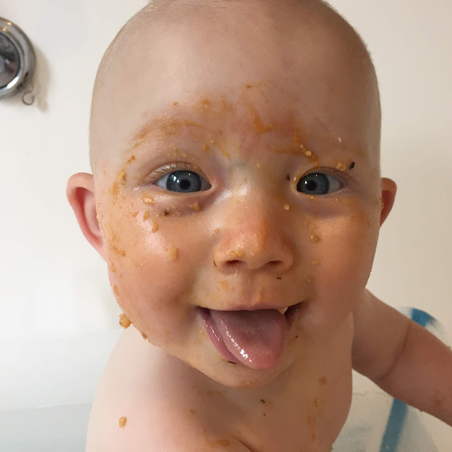 A baby sticking her tongue out with a face covered in food thanks to baby led weaning. She is sitting in the bath